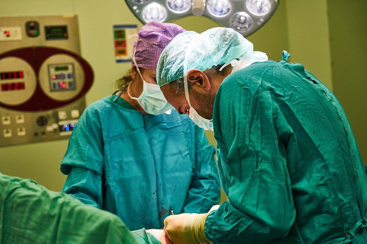Doctor wearing green scrubs performing surgery in operating room with nurse on opposite side wearing blue scrubs.