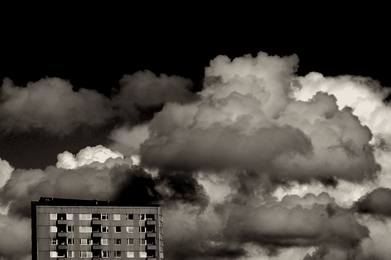 Black and white image of an apartment building in lower left corner and a dense cluster of assorted cumulus clouds behind with a black sky in the background