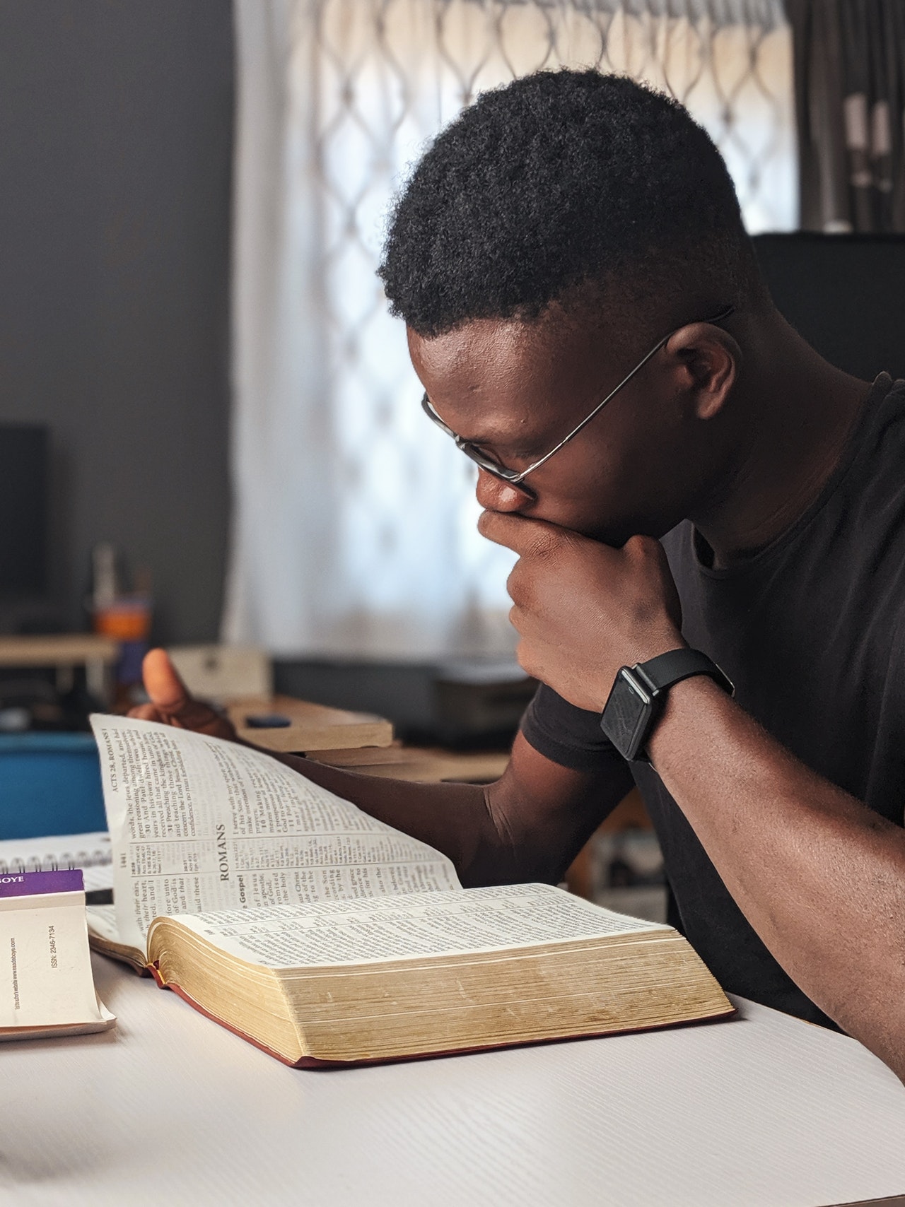 Photo of an African-American man wearing glasses while reading a Bible and holding his left up towards his mouth