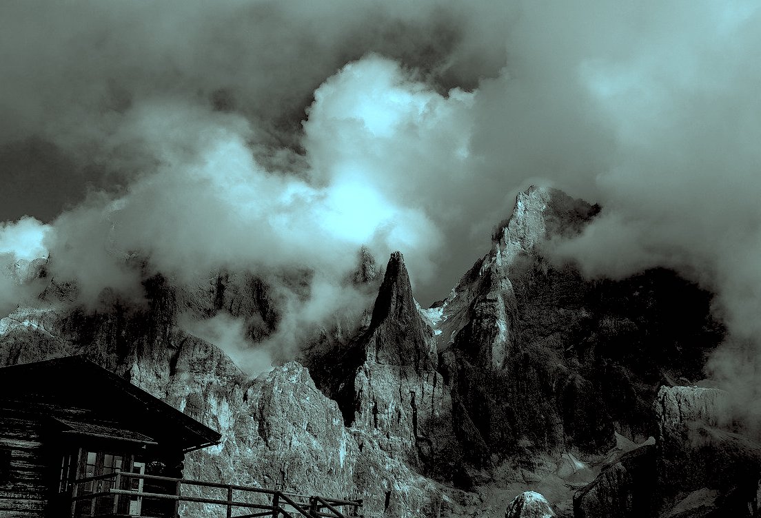 A blue tinted black and white image of a building in the left corner of the foreground with the pale di san martino mountain range in the background