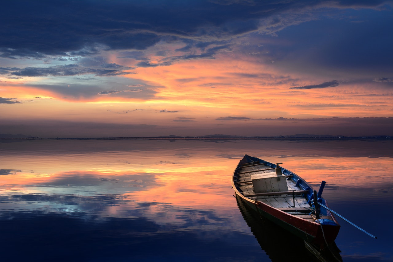 A multicolored sunset sky reflecting on a motionless sea in peace with a small wooden boat pointed outward in the lower right corner