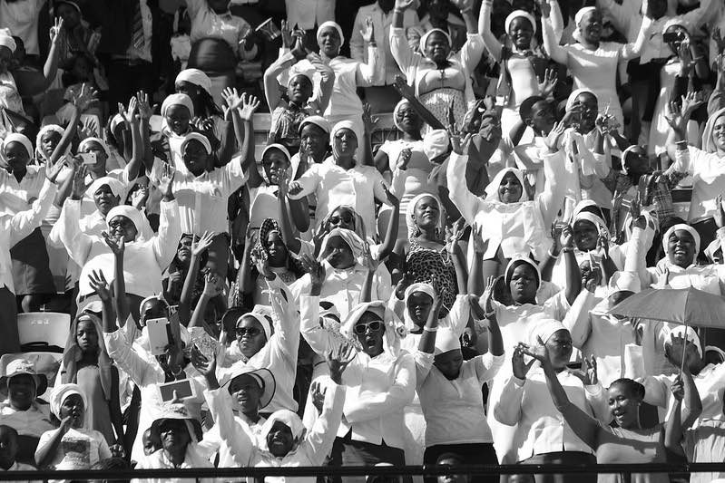 A group of African women in a stadium in the sun with some raising hands