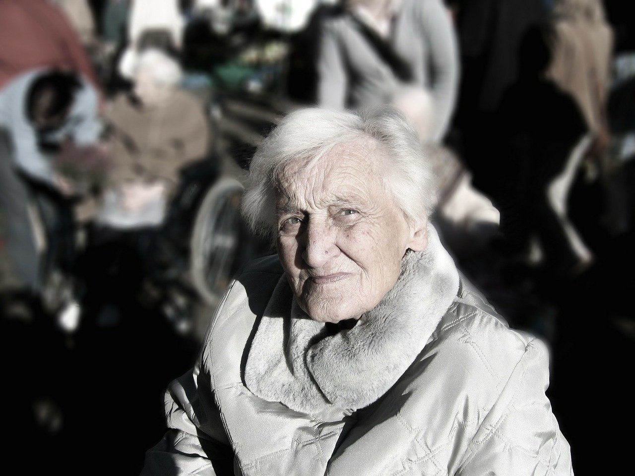 An older woman with white hair wearing a grey winter coat with bright sunlight on her face and looking suspiciously in the direction of the camera with many people in the distant background