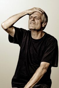 An older man seated with his right hand on his forehead and his eyes closed while looking slightly upward