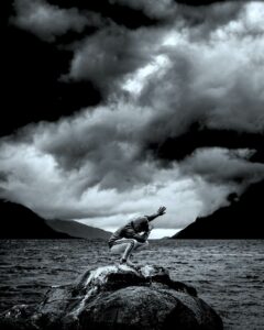 A young man in a hooded sweatshirt crouched on rocks in a 'dab' dance postion with ominous grey clouds, and water parting two mountains