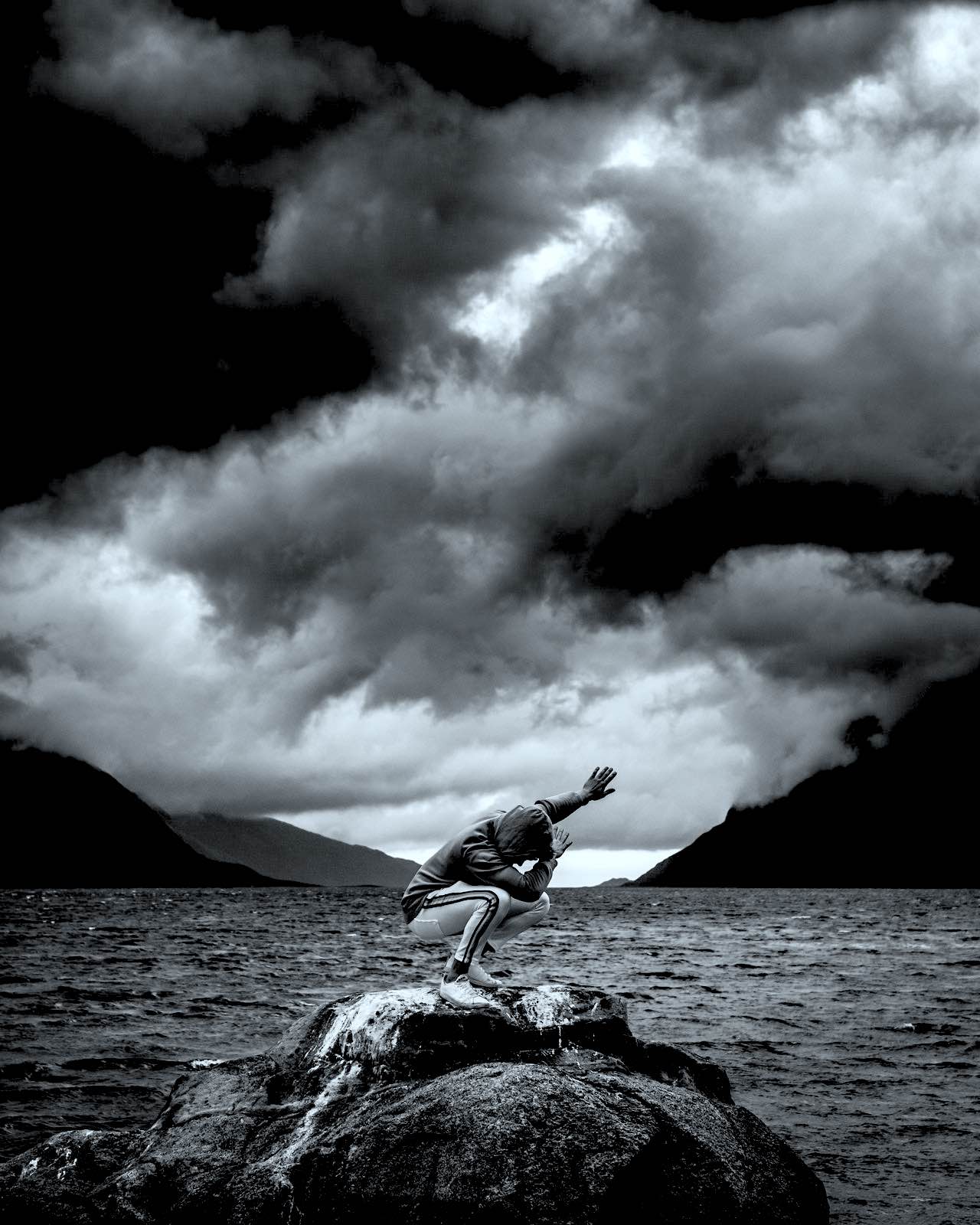A young man in a hooded sweatshirt crouched on rocks in a 'dab' dance postion with ominous grey clouds, and water parting two mountains
