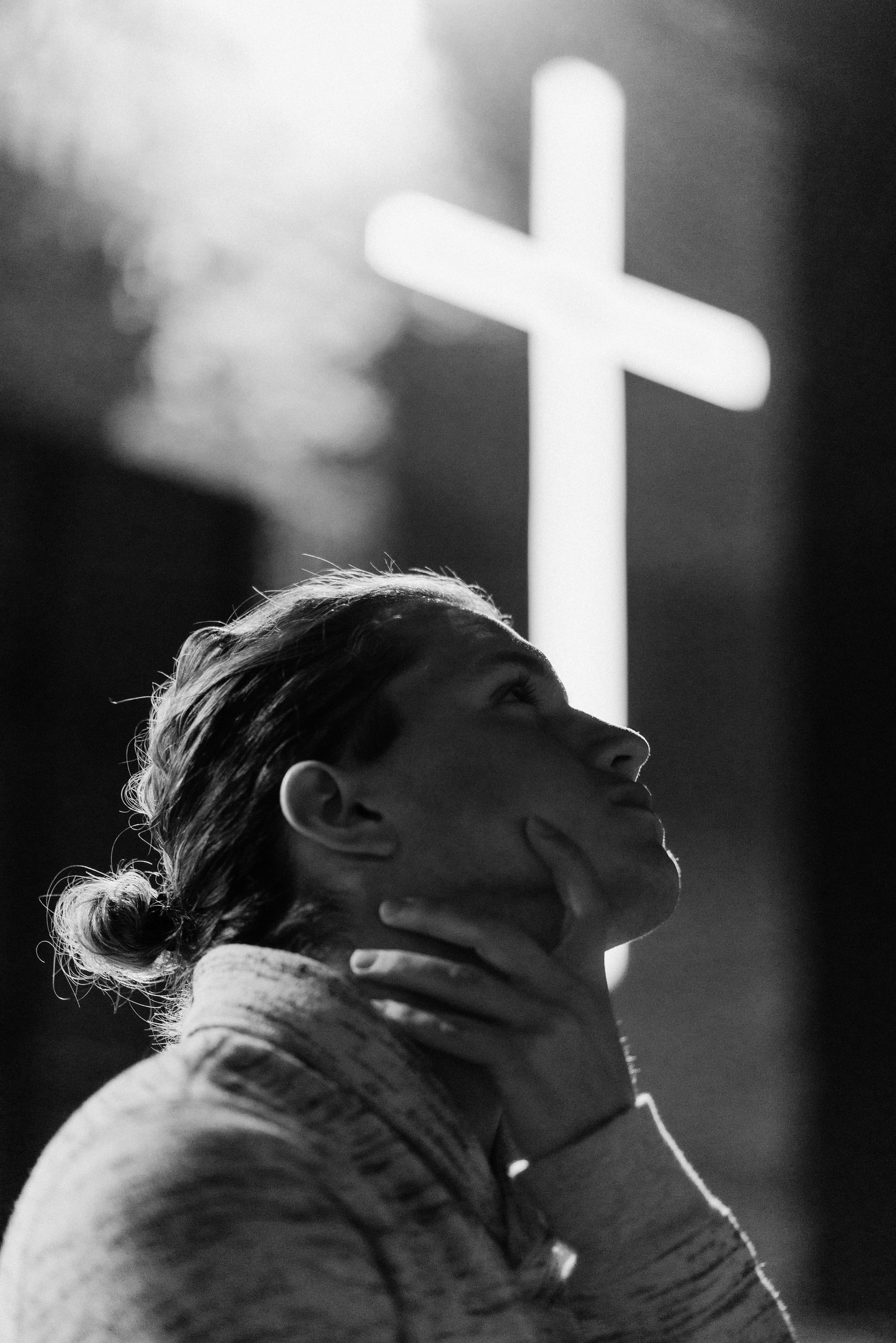A black and white photo of a woman with hand resting around neck looking up in contemplation with a blurred white cross in the background