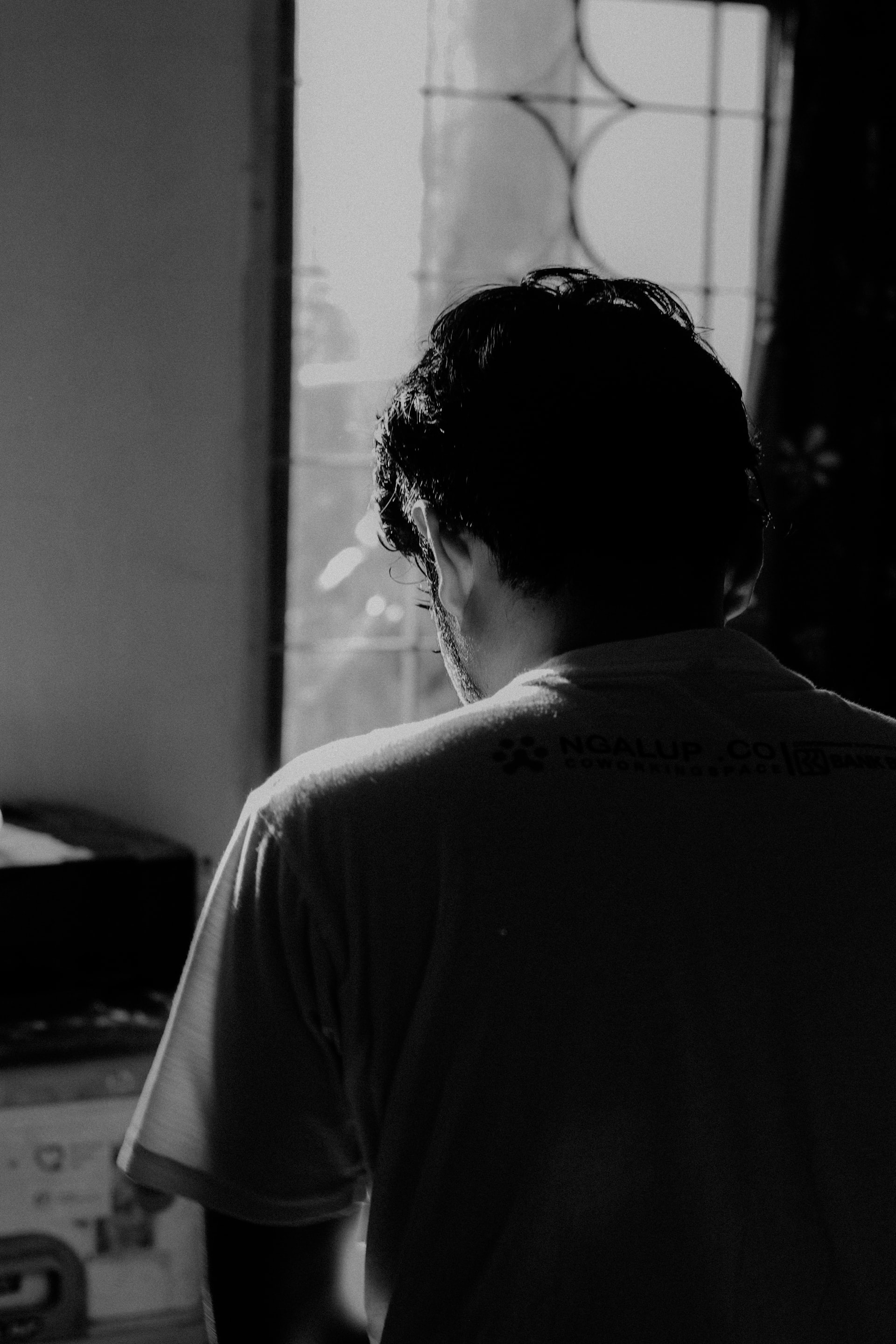 A black and white photo of the rear view of a man with his bowed down in a sun lit room