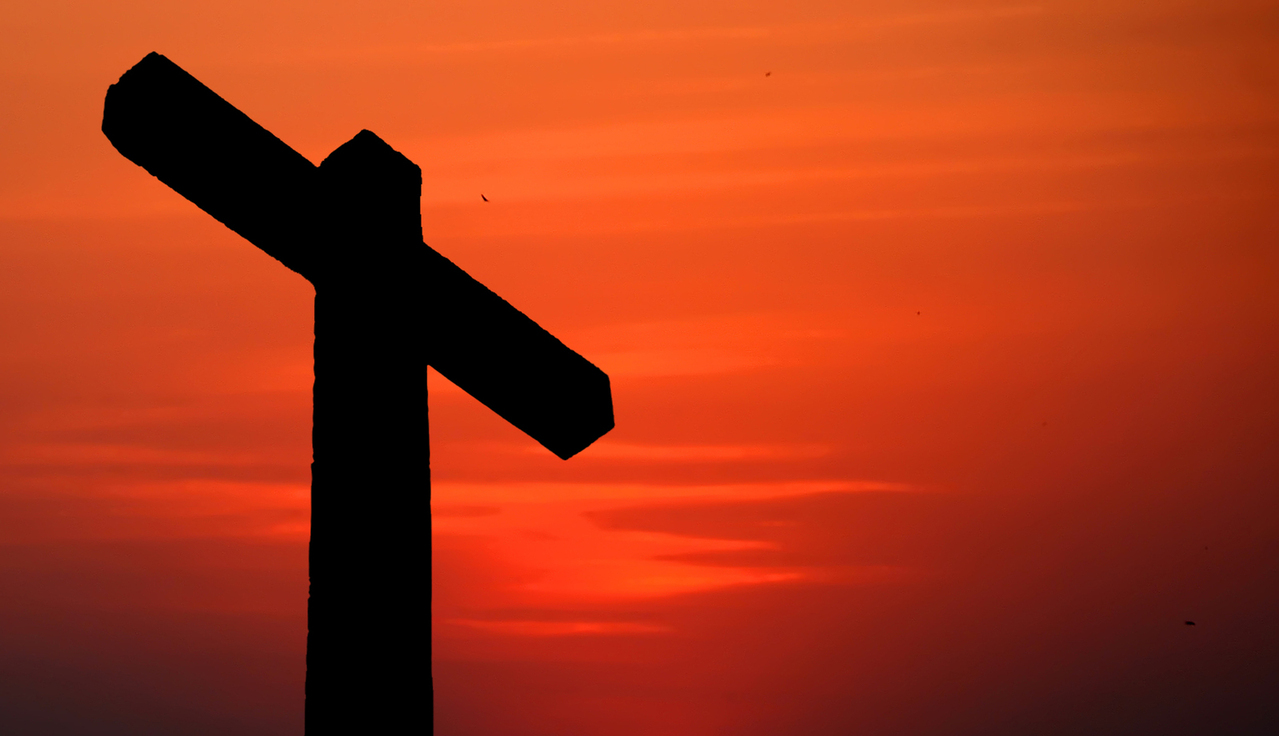 A silhouette of a cross on the left side of the frame with a dark reddish orange sky in the background