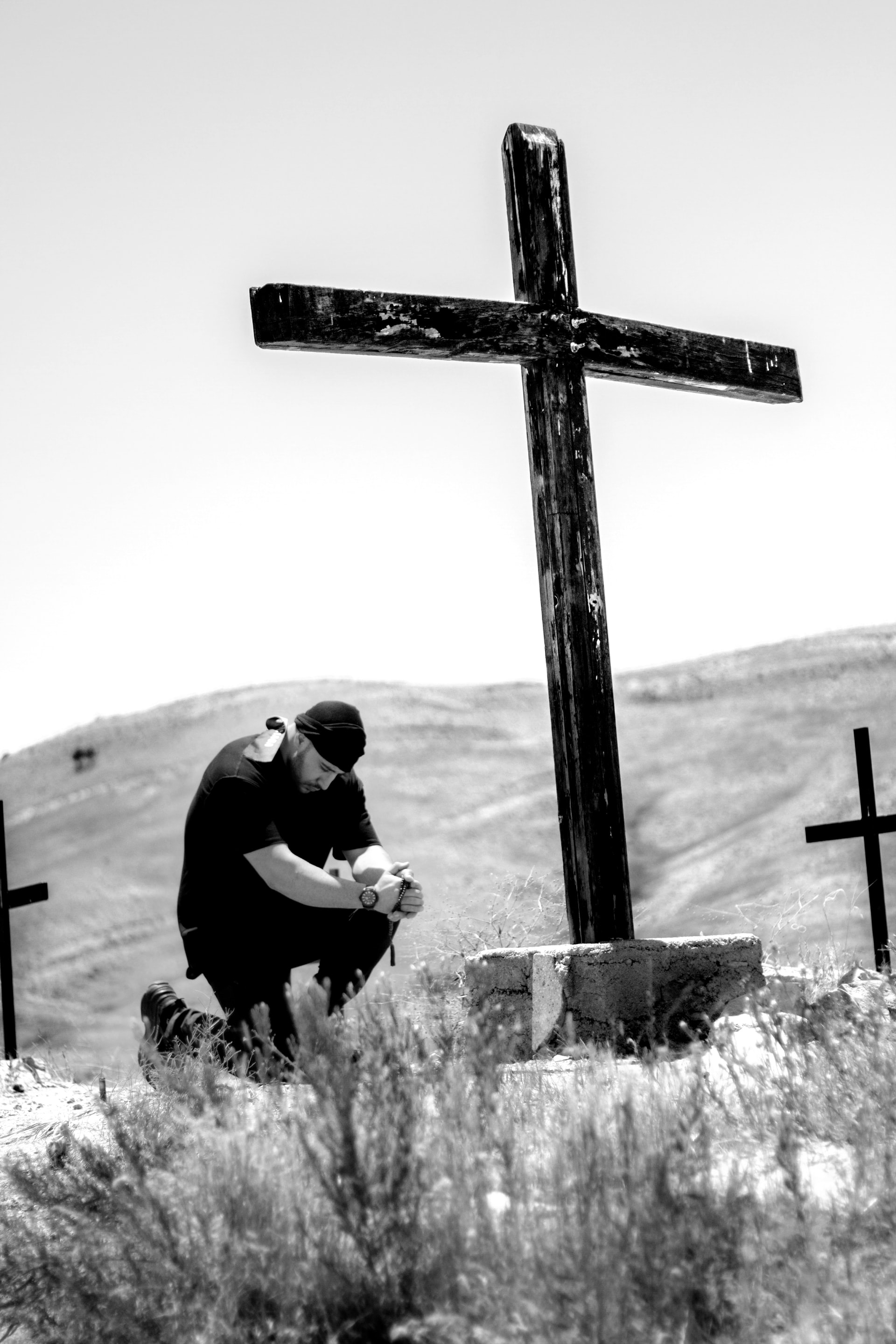 A black and white photo of a man dressed in black and wearing a black cap while kneeling at a large cross in a wilderness area and a large hill in the background