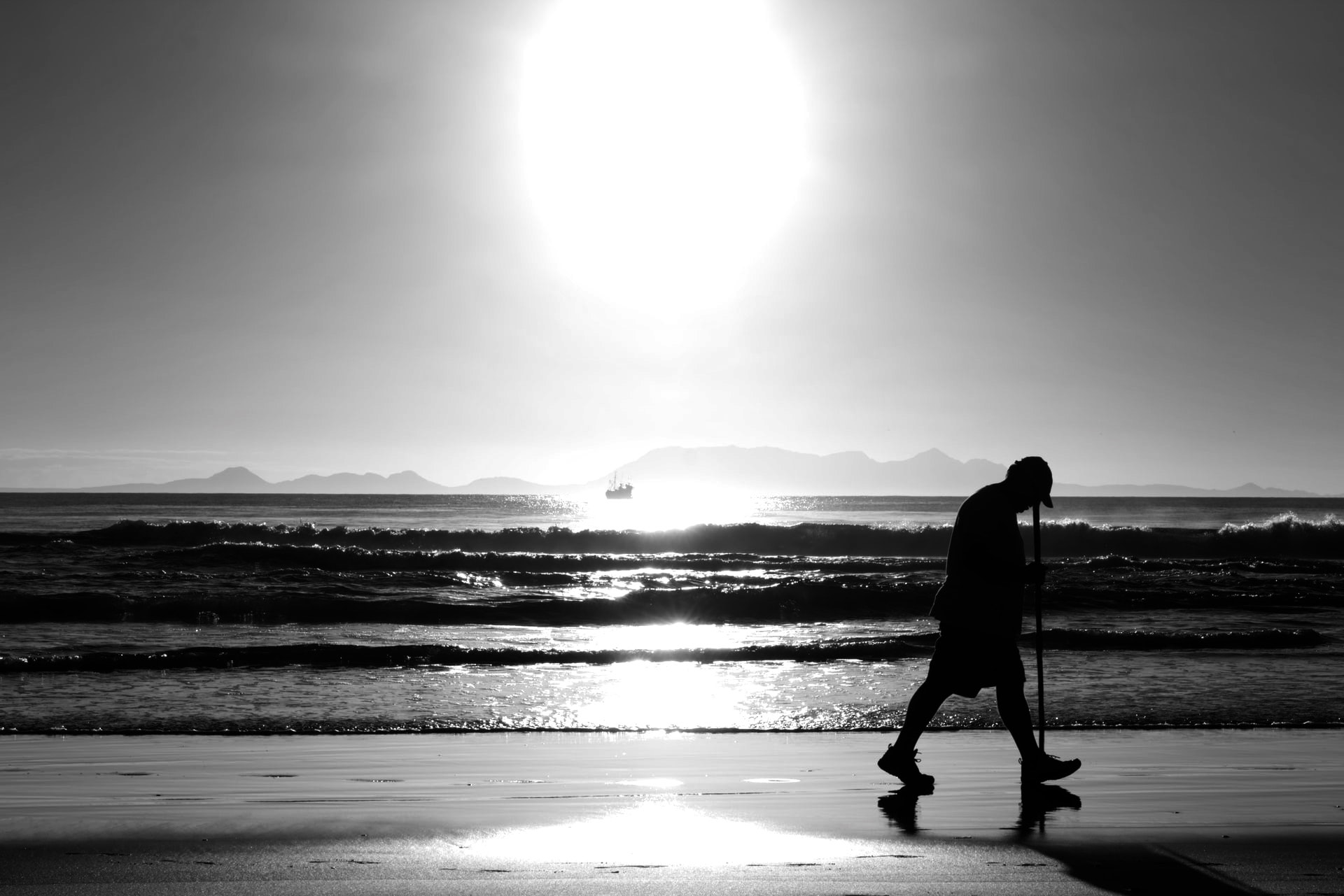 A black and white picture of a man walking solo, much like one who walked with God, on the beach with a bright sun near the horizon with a small vessel in the distance in the heart of the sun beam on the water