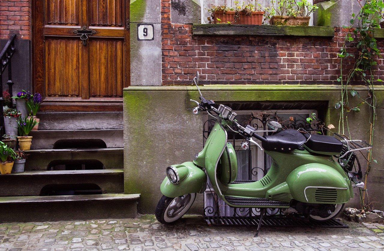 A photo of a green moped in front of the basement window of an old brick townhome symbolizing  a attempt to deny our self by scaling back our possessions