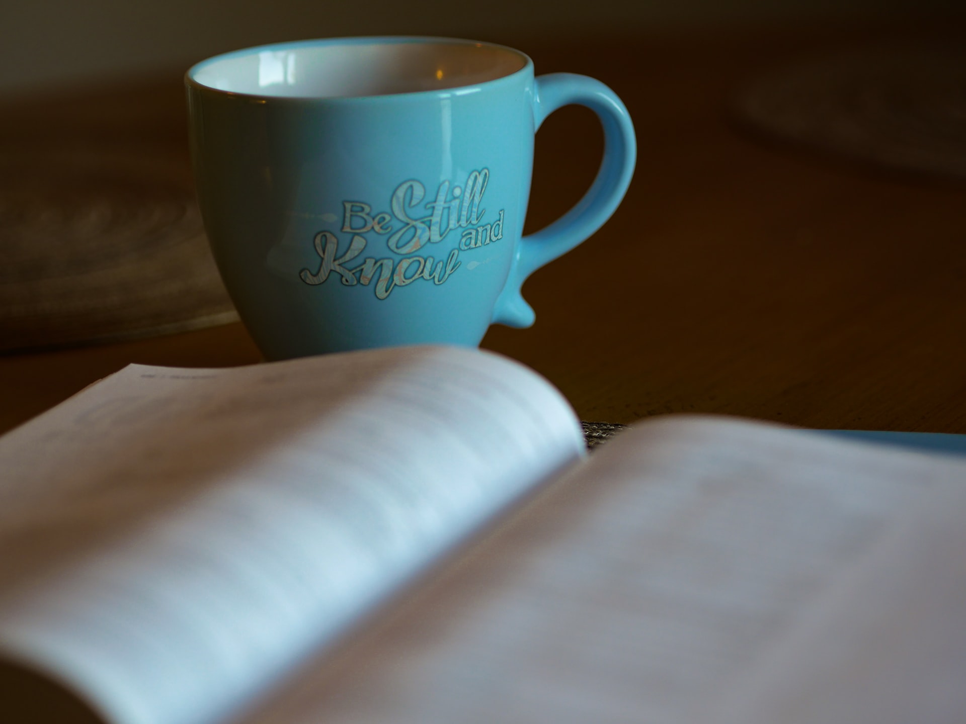 A photo of an open out of focus book lying flat on a table with a blue coffee cup immediately behind it