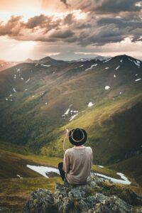 The backside of a man with a dark brown hat sitting on some rocks while looking out over grassy valley and hillside with clouds in the background