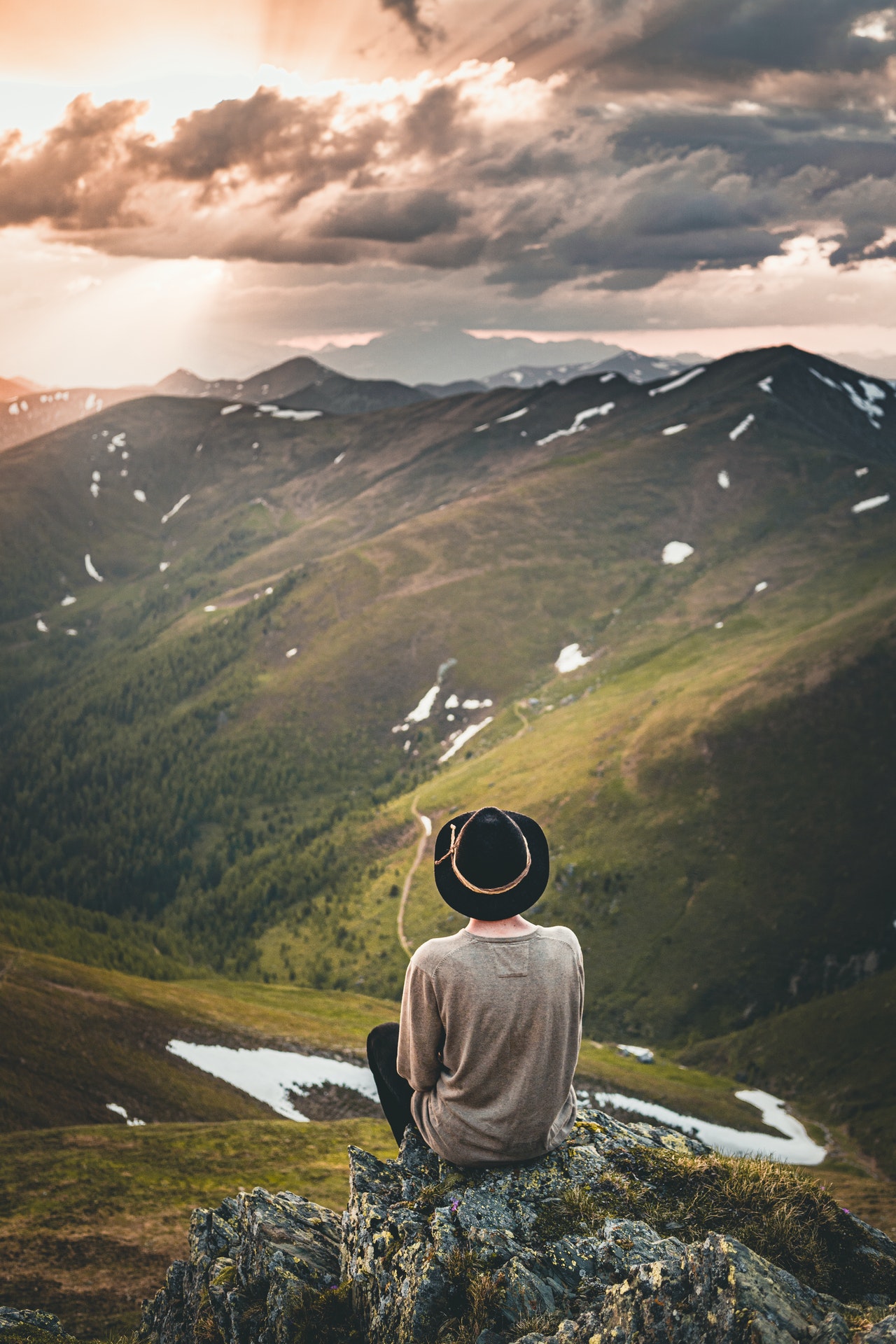 The backside of a man with a dark brown hat sitting on some rocks while looking out over grassy valley and hillside with clouds in the background