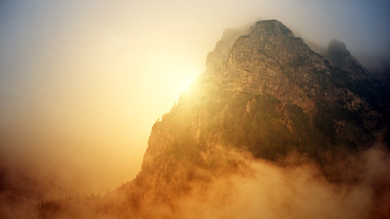 A large mountain with clouds below and a bright orange sun peering through the haze on the left side of it
