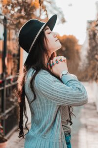 A young woman with a black hat and blue sweater standing an stone walkway with head held upward, eyes closed and hands folded together near her chin