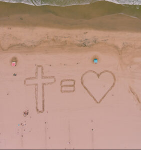 An aerial photo of a cross, an equal sign, and a heart written on a beach with the waves of greenish water crashing a short distance above them