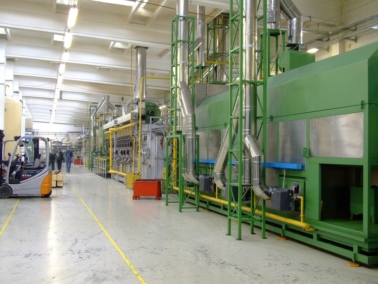 A factory hallway with large green and grey machinery on the right side, a forklift on the left, and several workers walking in the distance