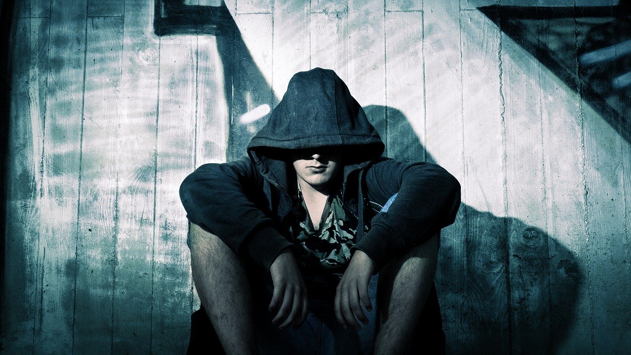 A blue tinted photo of a young caucasian man with arms resting on his knees seated against a white fence with some graffiti, while wearing a hoodie with the hood over most of his head