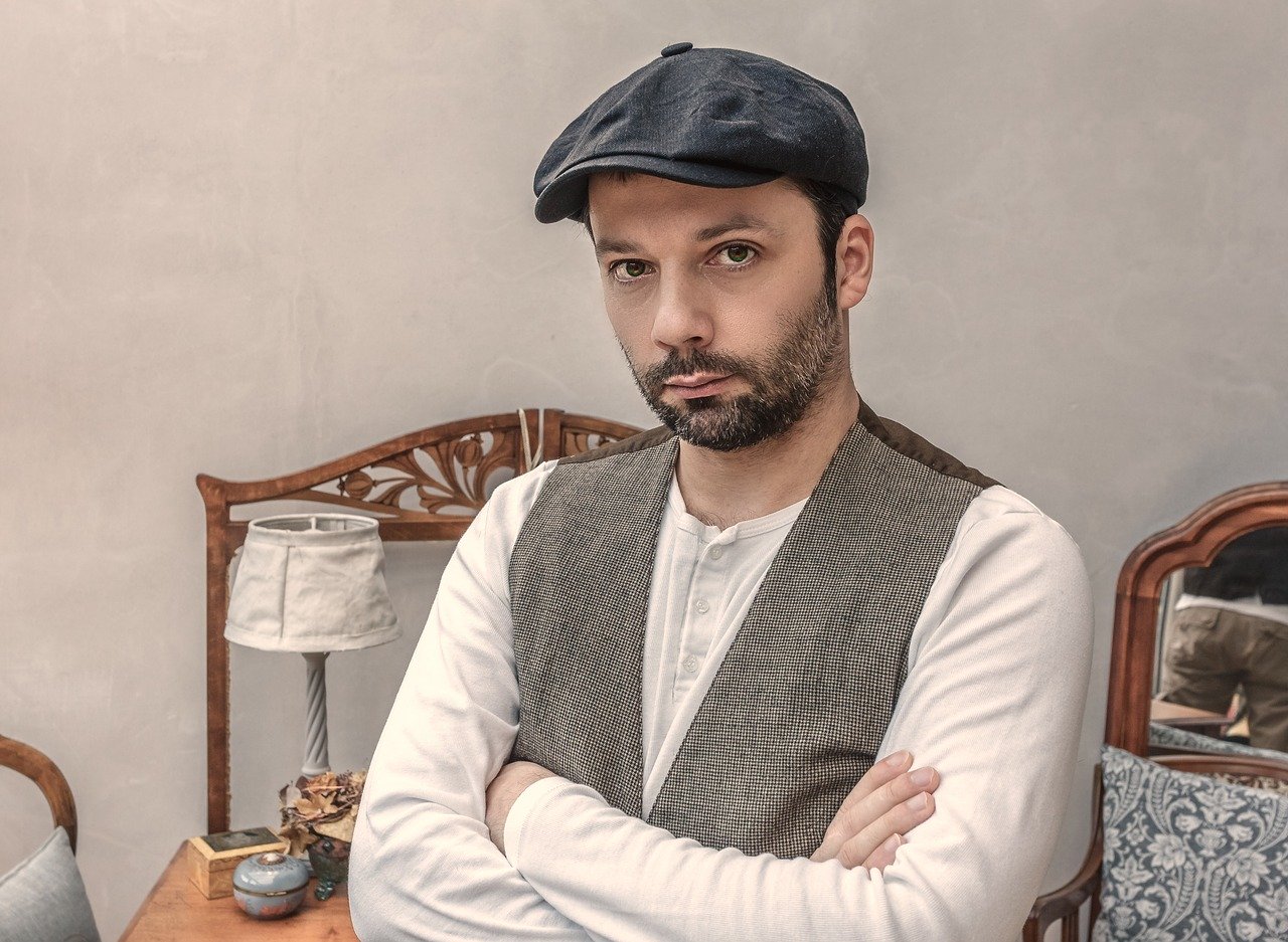A man with a thin beard and mustache seated in a bedroom with his arms folded and wearing a white long sleeve shirt, grey vest, and a dark blue cap with a stern facial expression possibly complaining