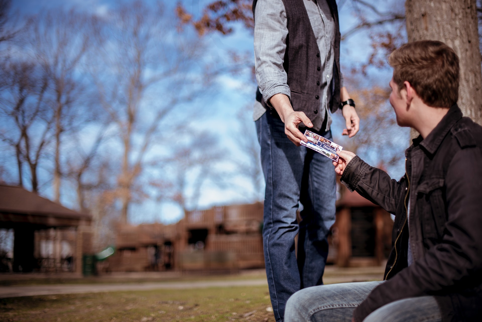 A person standing wearing jeans, grey shirt, and a dark vest handing literature and demonstrating the love of God to a young caucasian man with brown hair, dark jacket, and grey jeans seated at a tree in a courtyard of a residential area with a blue sky in the background