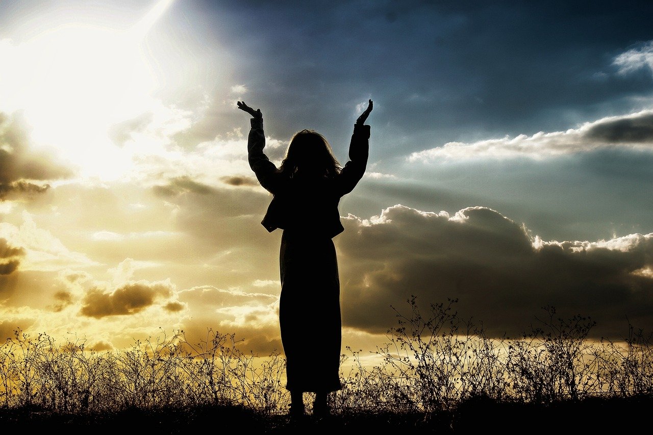A silhouette of a woman wearing a skirt standing among grasses with arms raised and sun poring through assorted clouds in a pale orange and blue sky