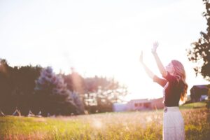 A photo of a woman on the right side of the picture with arms raised, eyes closed, and standing over a grassy field with trees and farm buildings in the distance and a bright sun overhead