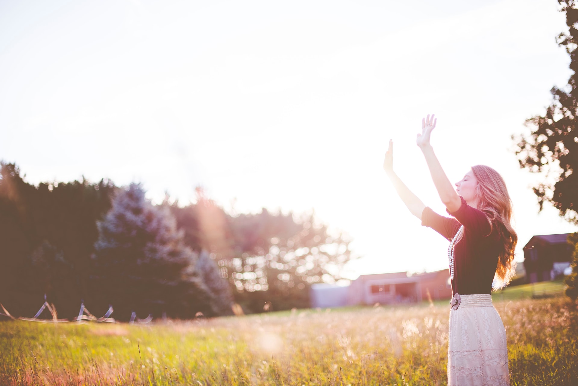 A photo of a woman on the right side of the picture with arms raised, eyes closed, and standing over a grassy field with trees and farm buildings in the distance and a bright sun overhead