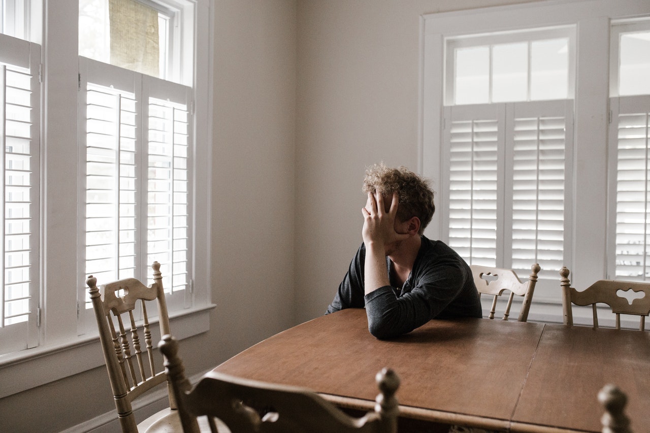 A caucasian man with bushy brown hair sits at the corner of a table in a room with sunlight poring through louvered blinds with head resting in left hand baffled over some wrongdoing