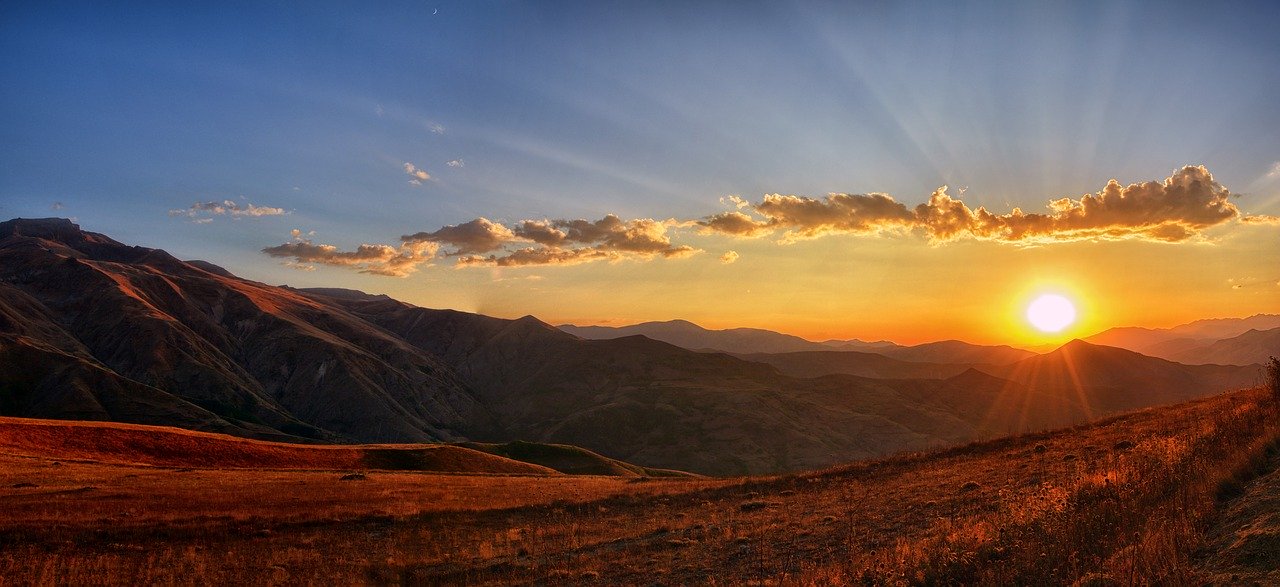 A view of a sunrise from a mountain plateau with other mountains in the background and a thin row of clouds in the blue and orange sky