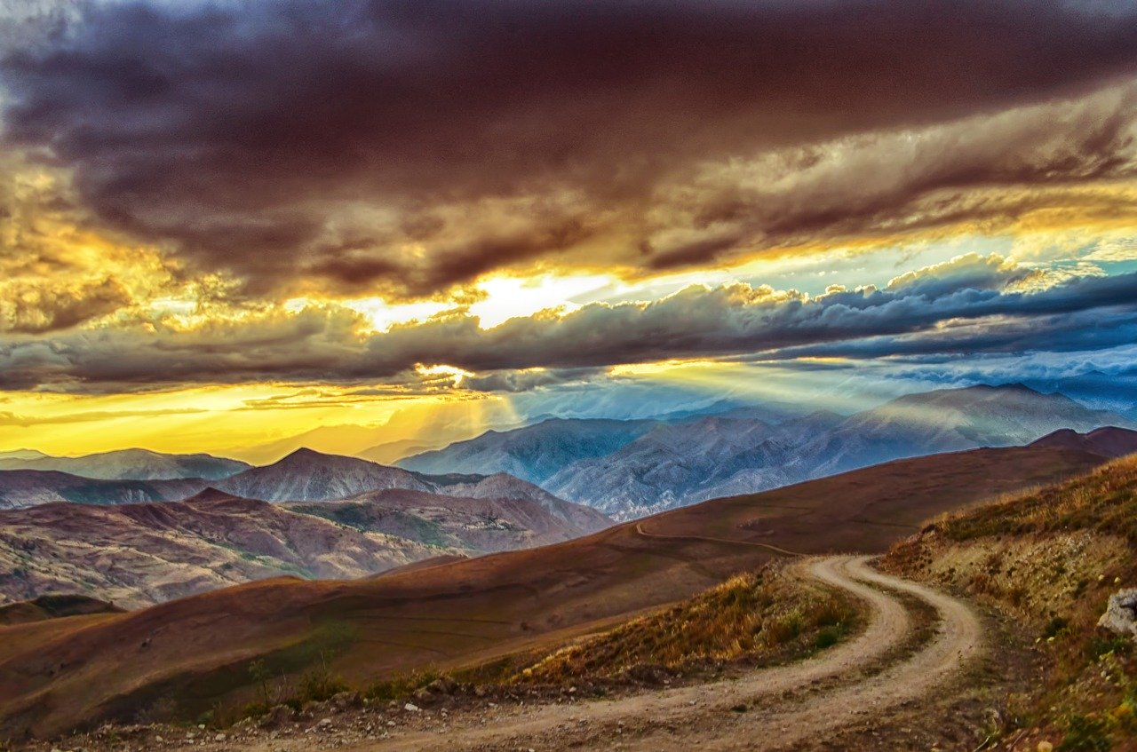 A winding dirt road leading out of a series of brown rolling hills and grey mountains with a mixture of grey/brown, blue and yellow clouds and sky overhead along with beams of sunlight poring through