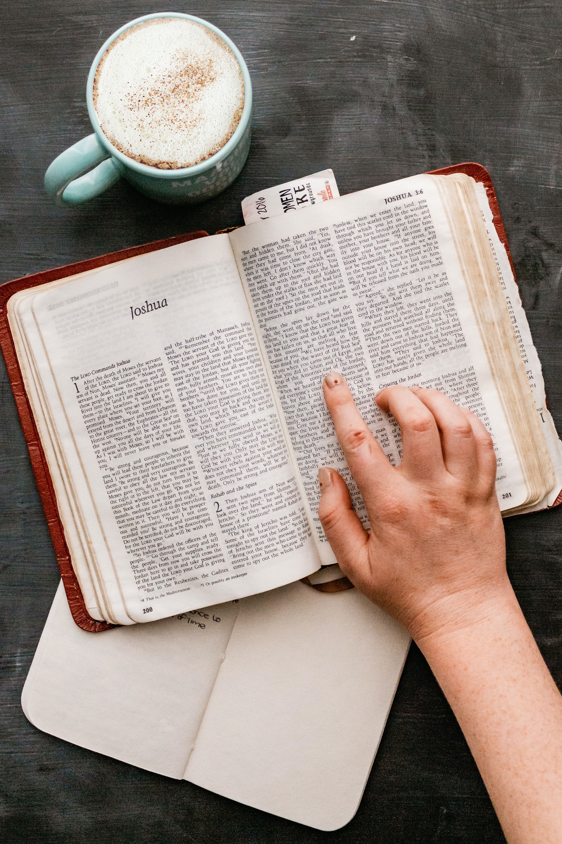 A Bible laying flat on a dark gray surface, opened to the book of Joshua with a hand and index finger pointing to a verse, and a green mug filled with latte rests nearby
