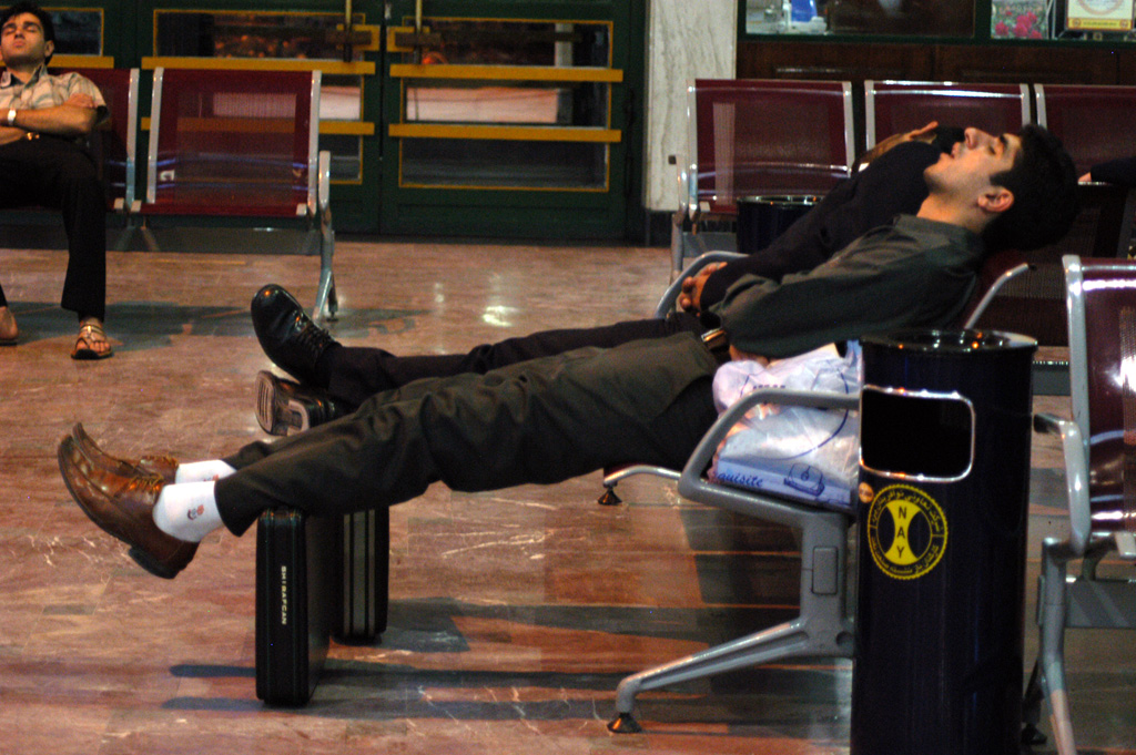 A caucasian man lying in a chair, asleep, in an airport terminal with his legs propped up by a piece of luggage, showing the need to stop sleeping 