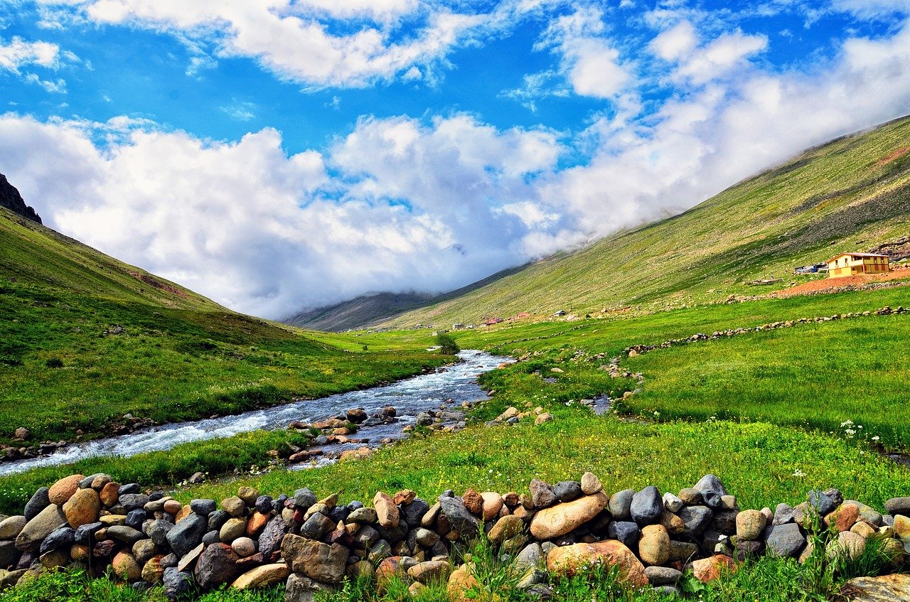 A small river leads out between two green grassy mountainsides with a yellow home in the distant right, large stones in the front, and clouds with a blue sky in the background