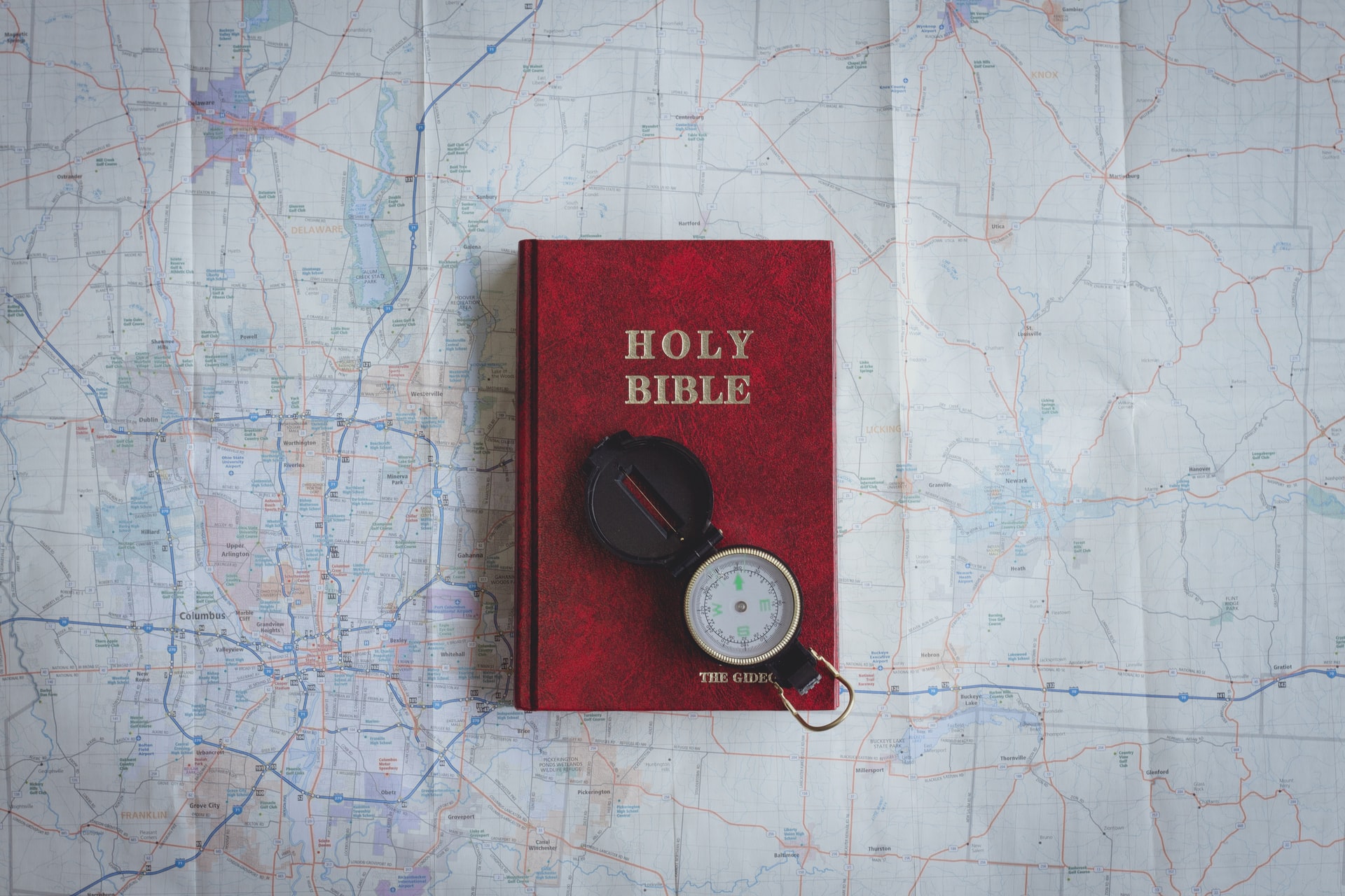 An overhead view of a red Bible, a key to victory, with a compass resting on top in the lower right corner, both are laying on top of a large road map 