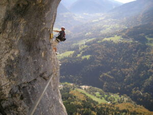 A view from the side of a mountain of a young man scaling the same mountain on a sunny day with tree filled hills and mountains in the background