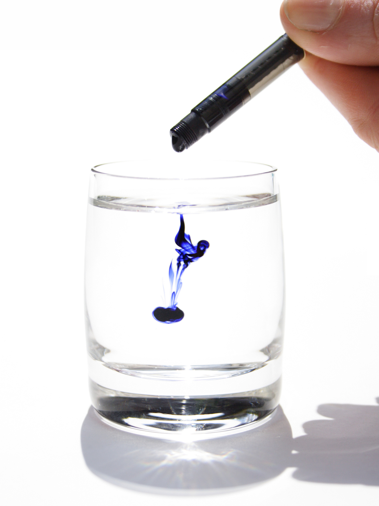 A short clear glass of water with a small amount of blue ink dispersing inside from a vial held overhead in a brightly lit space