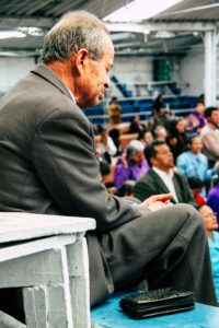 An older Hispanic/Asian man seated by himself with a group of men and women in the background in a small auditorium or gymnasium with eyes closed and head bowed in prayer