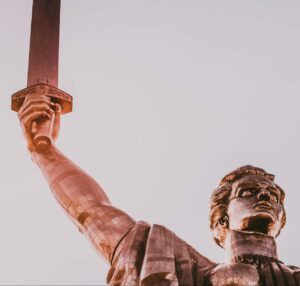 A partial bust of a metal statue depicting a man holding in the air a sword and shield on a grey-white background in victory
