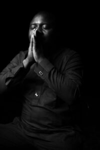 A black and white photo of an African man seated with hands raised and pressed flat together against his mouth and nose and eyes closed