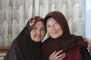 A photographic portrait of two older Asian woman wearing hooded garb with one partially embracing the other in front of a pleated white patterned curtain