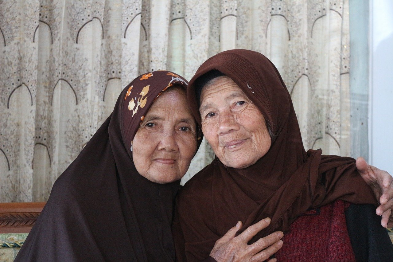 A photographic portrait of two older Asian woman wearing hooded garb with one partially embracing the other in front of a pleated white patterned curtain
