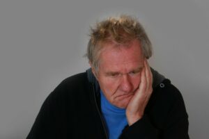 A older caucasian man wearing a black jacket and blue t-shirt seated with his left hand pressed against the side of his face in a sad, disturbed expression