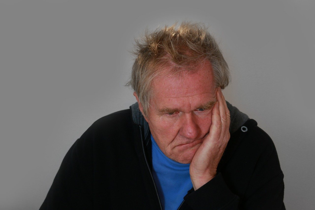 A older caucasian man wearing a black jacket and blue t-shirt seated with his left hand pressed against the side of his face in a sad, disturbed expression