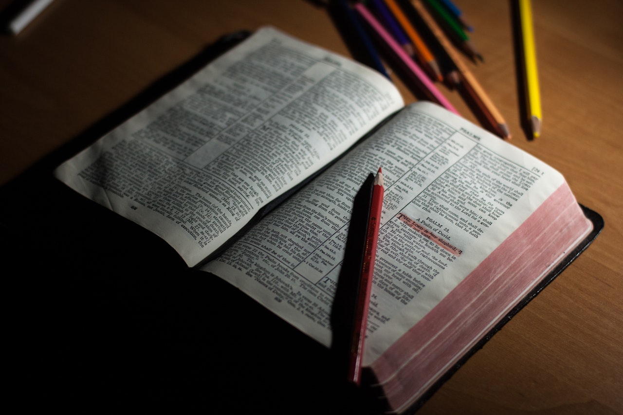 An open Bible laying flat on a table in low light with a red colored pencil resting on one half and several other colored pencils nearby