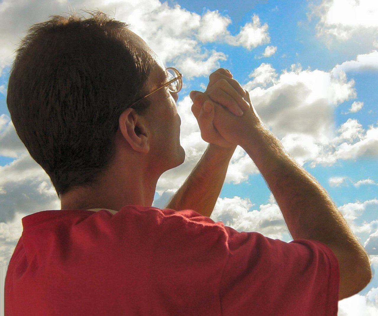 The partial back side of a Caucasian man with dark hair, glasses, and a red short sleeved shirt holding his clinched hands together in the air towards a the sun and a partially cloudy sky