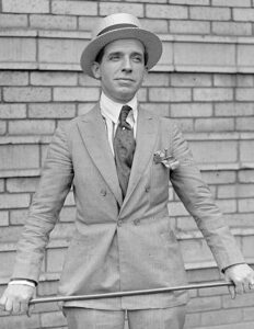 A black and white photographic portrait of Charles Ponzi standing against a brick wall holding a bar at waist level