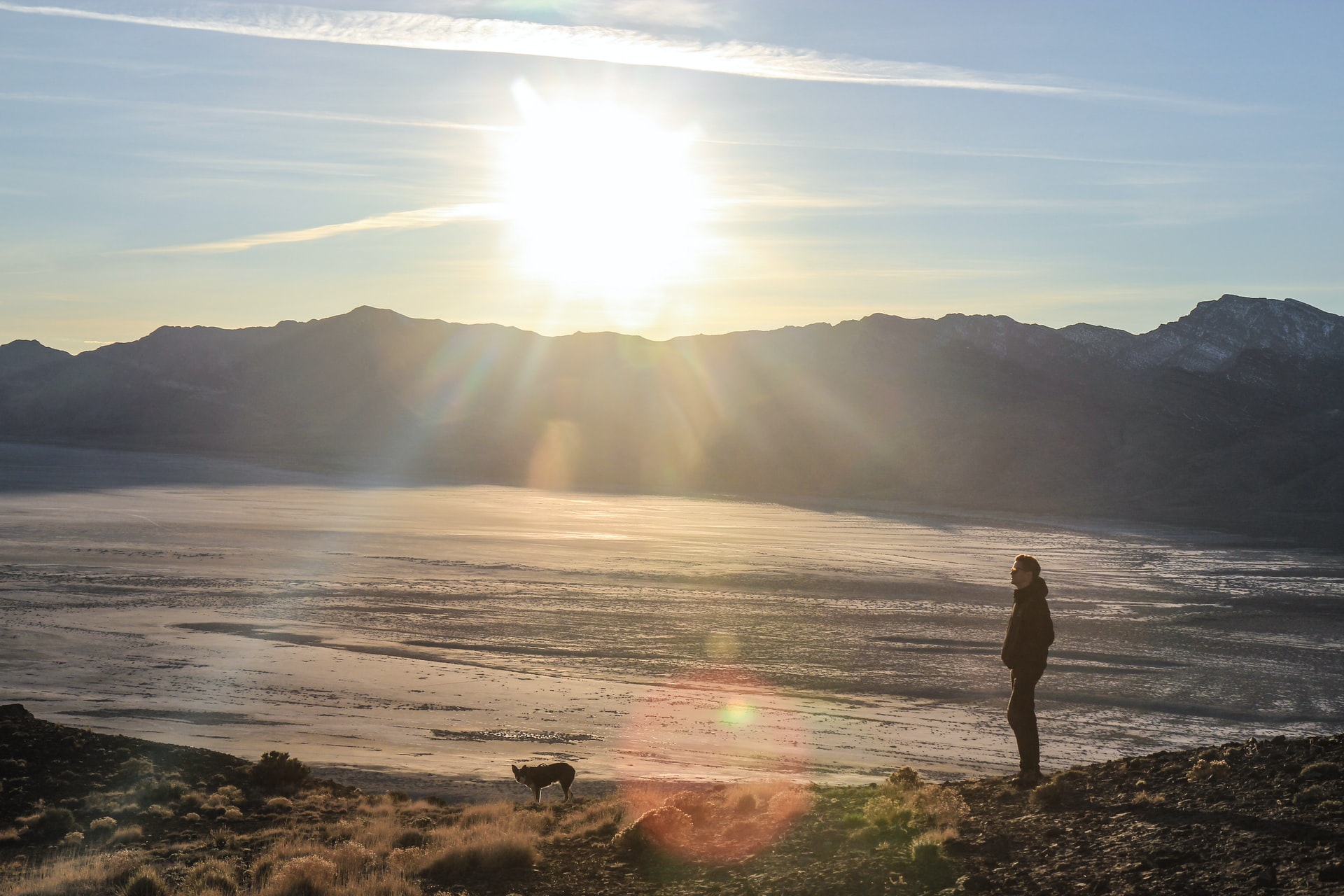 A man with a small dog standing on the shore of a lake with mountains, a bright sun, and blue sky in the background