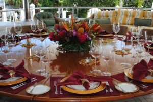 A large round table set with elegant glassware, fine china, and a colorful floral centerpiece in a covered outdoor dining area with outdoor furniture in the background